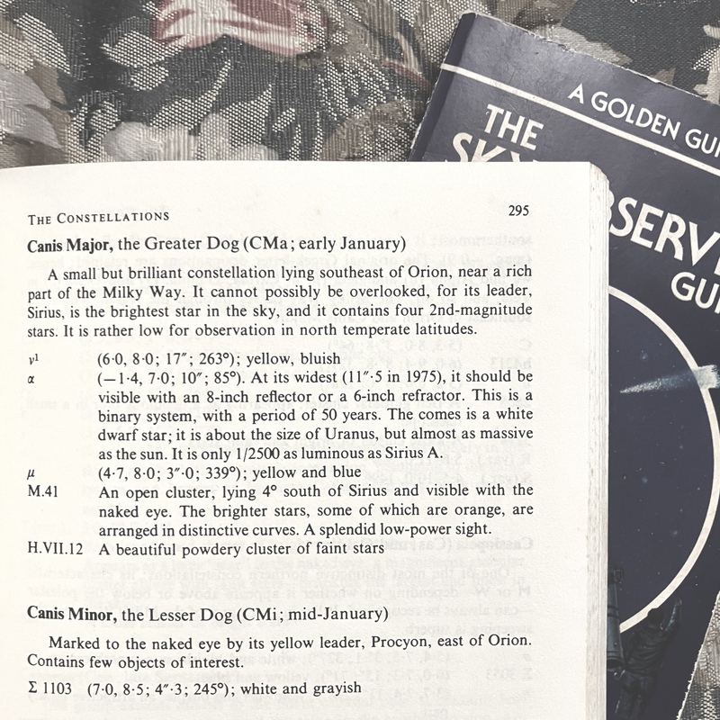 a photo of two books on a floral fabric background. "the sky observer's guide" is underneath, and on top is "the amateur astronomer's handbook" opened to page 295. at the top it reads "the constellations" and has information about canis major and canis minor.