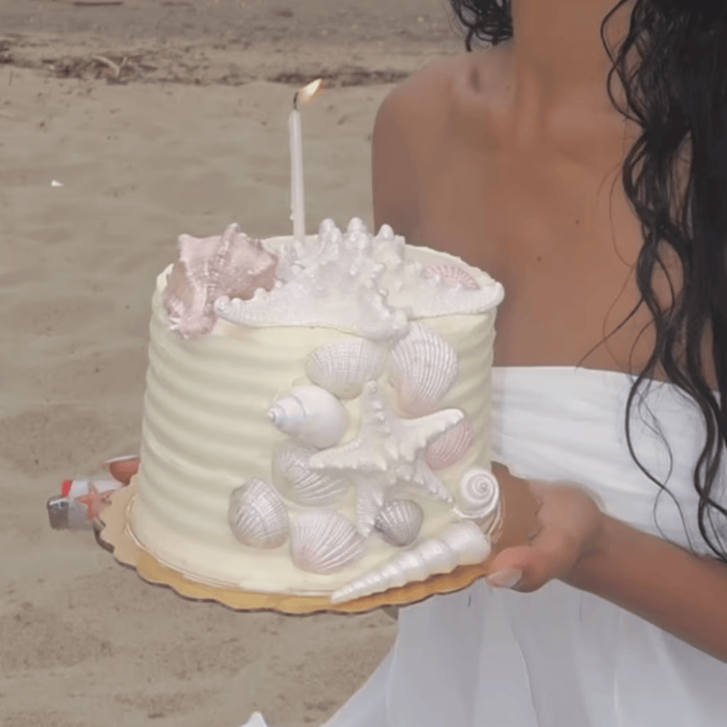 a cropped photo of a person on a sandy beach holding a birthday cake and lighter. the cake is white with a lit candle and iridescent white and pink seashell decorations.