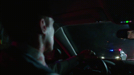 a gif from the show of adrian driving with police cars passing him in the background. he is covered in blood spatter and starting to smile, lick his lips, then whistle.